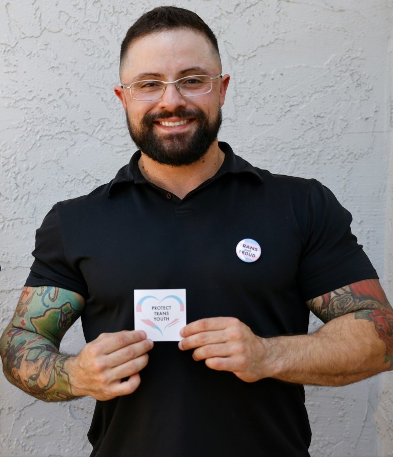 a man in a black polo shirt standing in front of a white outdoor wall, holding a sticker that depicts hands making a heart. the sticker is colored with the colors of the transgender flag