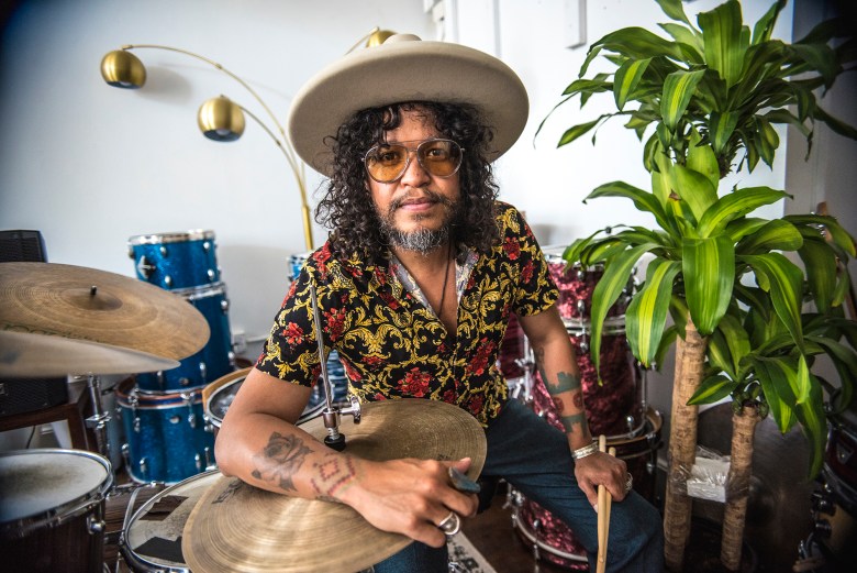 Daniel Villarreal sits among his impressive collection of drums, with his forearm draped across a hi-hat and a pair of drumsticks in his other hand.