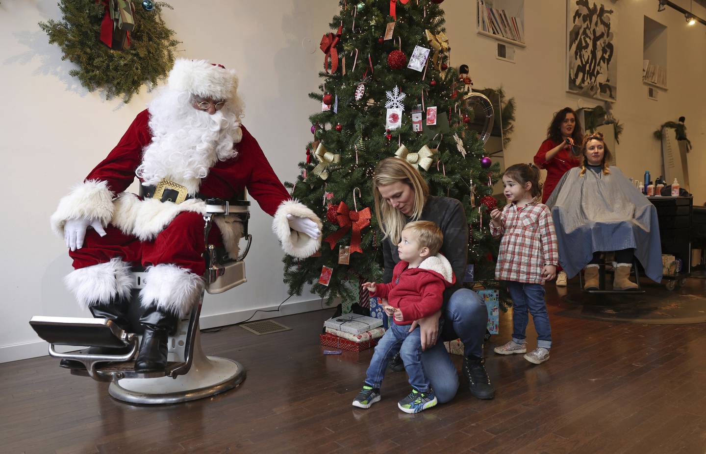 Ronnie Trejo, portraying Santa Claus, sits in a barber's chair as Kristie Stewart encourages her son, Graeme, 1, to take pictures with him at YO:U hair salon on Dec. 10, 2022, in Chicago. Trejo said he has played Santa for more than 20 years, in more recent years at the salon, which is owned by his daughter, Yolanda Trejo, second from right. 