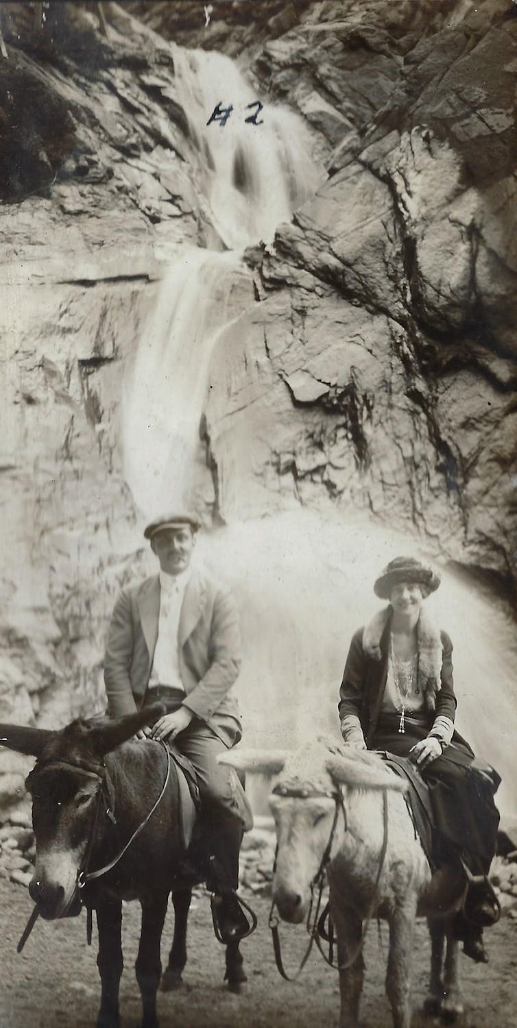 A white man and woman are seen riding on donkeys near a mountain waterfall.