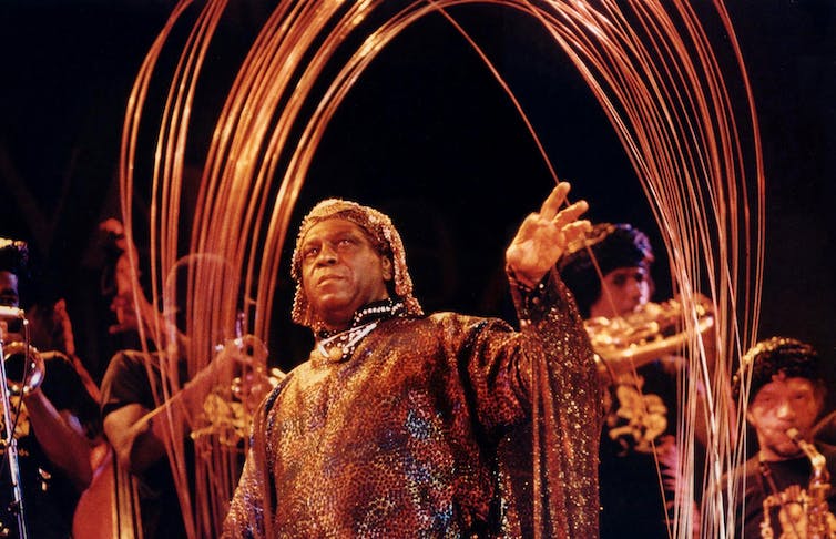 A man in an elaborate headdress and cloak stands in front of a steel structure.