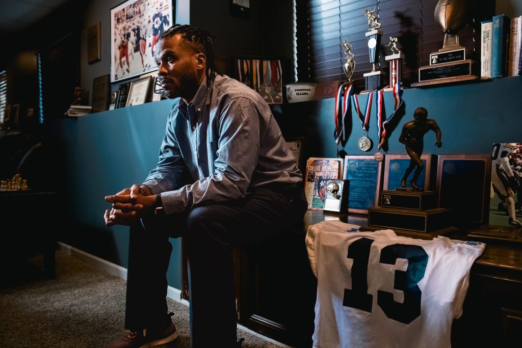 Johnny Johnson, Jr., former NFL Detroit Lions quarterback, in his home surrounded by his #13 collegiate jersey, trophies, medals, and other memorabilia of his past career.