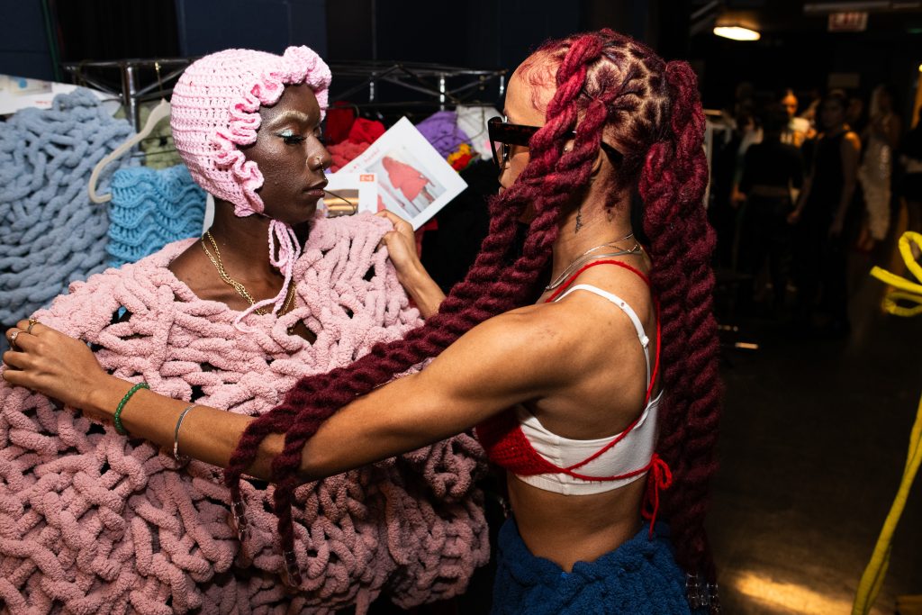 Chelsea B, a streetwear fashion designer, is helping one of her models get dressed in her design behind the stage.