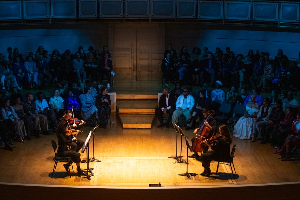 String quartets play classical music on stage at The Chicago Symphony Orchestra Hall. In the background is an audience listening.