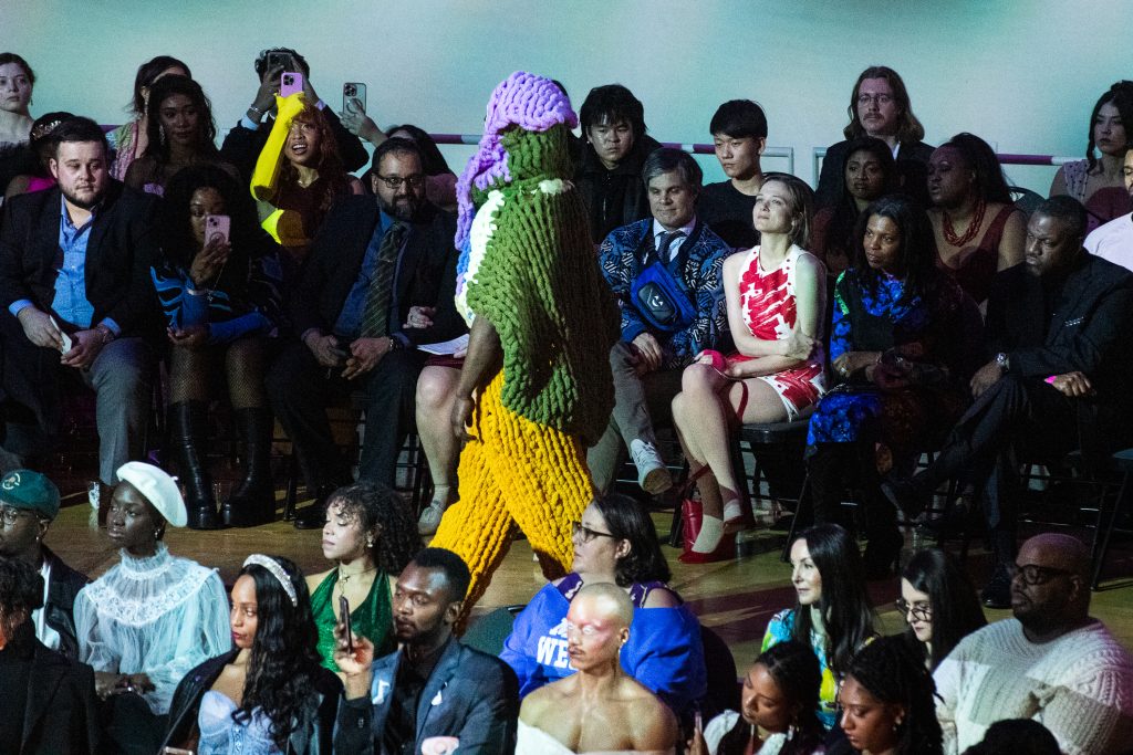 A fashion model is walking down the runway wearing fashion designs by Chelsea B, a celebrity streetwear designer. In the background are guests of the event watching the model walk.